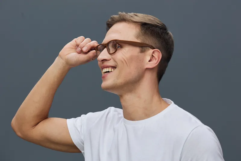 Smiling young man with glasses adjusting his eyewear, casual white t-shirt, cheerful expression, friendly appearance, outdoor setting, natural light, positive vibe.