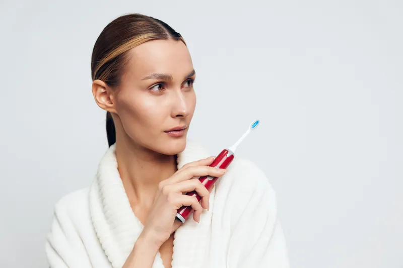 A young woman in a white bathrobe holds a red electric toothbrush near her face, looking thoughtfully to the side against a plain white background. Concept for dental care and hygiene.