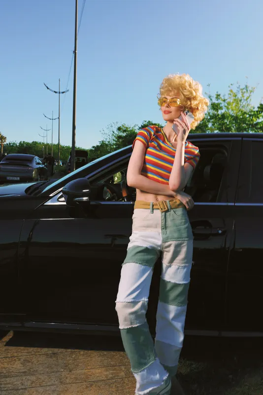 Young woman with curly blonde hair leans on a black car in a sunny parking lot, wearing a colorful striped top and patchwork jeans, exuding retro style and confident mood.