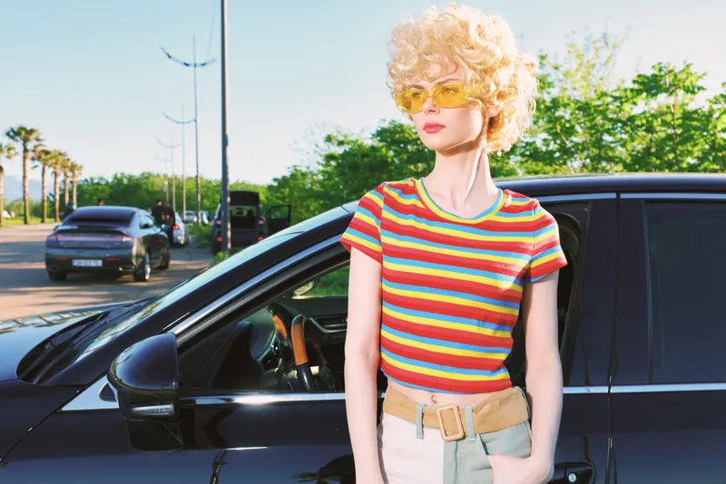 Young woman with curly blonde hair stands beside a dark car wearing a bright striped cropped tee and light denim shorts, exuding retro film color mood and confident casual style.