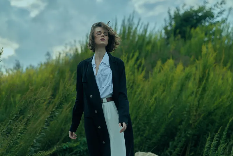 Fashion, outdoor, coat, dress, wind, confidence, woman standing in tall grasses wearing a dark long coat over a white dress, breeze lifting fabric for a dramatic, stylish look