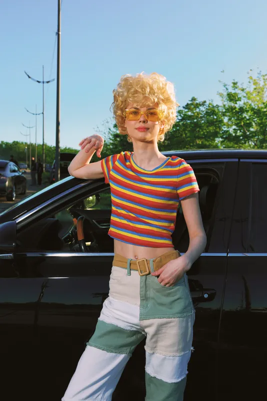 Smiling woman with curly blonde hair wearing a striped crop top and patchwork pants by a car in bright daylight. Retro film color vibe, lifestyle and fashion mood.