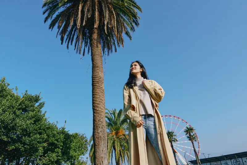 Outdoor palm scene with a stylish woman in a long coat under a clear blue sky, showcasing fashion sense and confident pose beside a park and distant ferris wheel