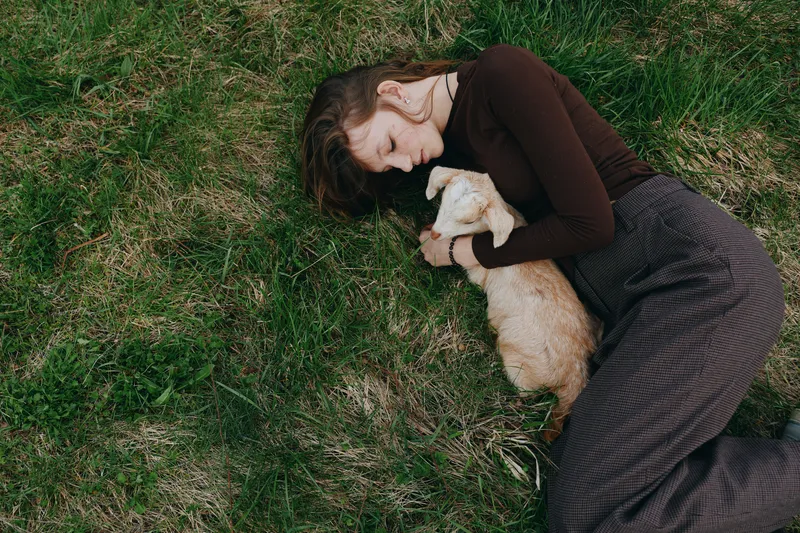 Woman lying on green grass, embracing a baby goat in a tender moment. Outdoor scene with natural light, relaxed mood, gentle bond between human and animal, rustic calm, simple lifestyle