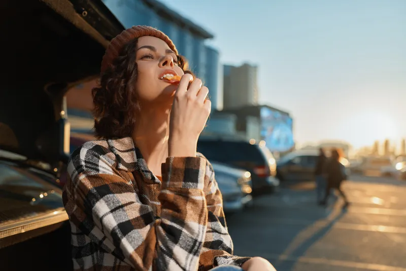 Woman eating snack in beanie and plaid shirt at roadside during sunset, candid lifestyle portrait with golden hour glow, authenticity, mindful living and emotional storytelling.