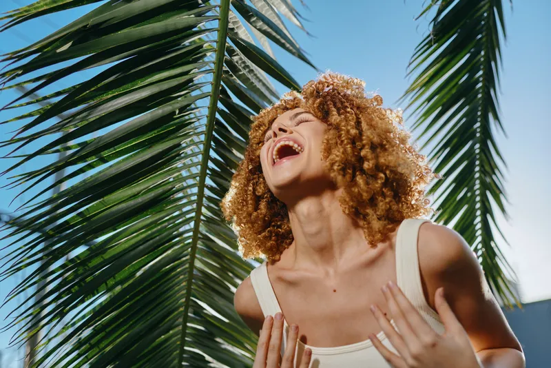 Laughing woman with curly hair enjoying sunny tropical outdoors near palm leaves, dressed in white tank top under clear blue sky with bright sunlight.