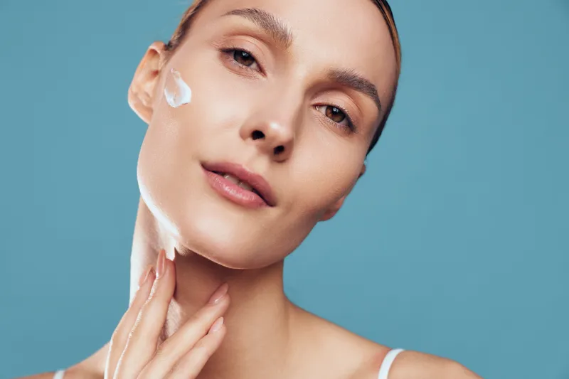 Woman applying face cream in skincare, beauty and spa wellness, white texture on cheeks, soft studio light, blue background, self care concept, calm mood, now