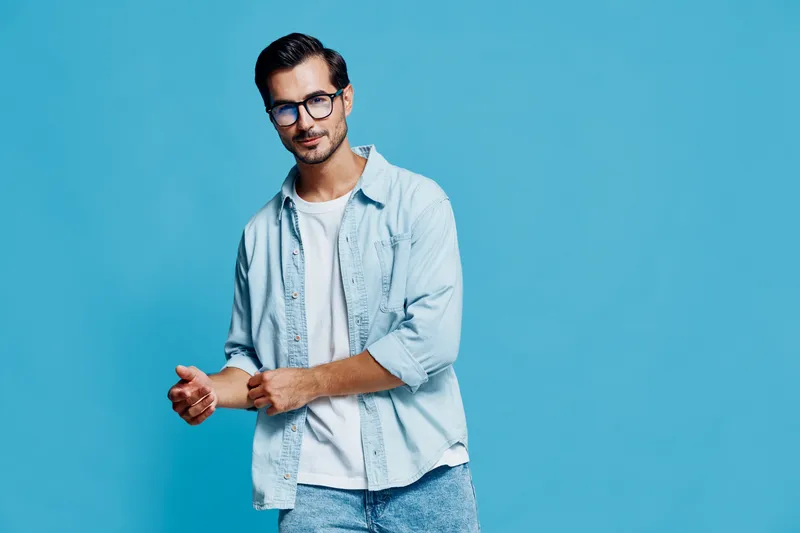 Stylish handsome young man with glasses posing confidently in light denim attire against a vibrant blue background, showcasing a modern lifestyle concept.