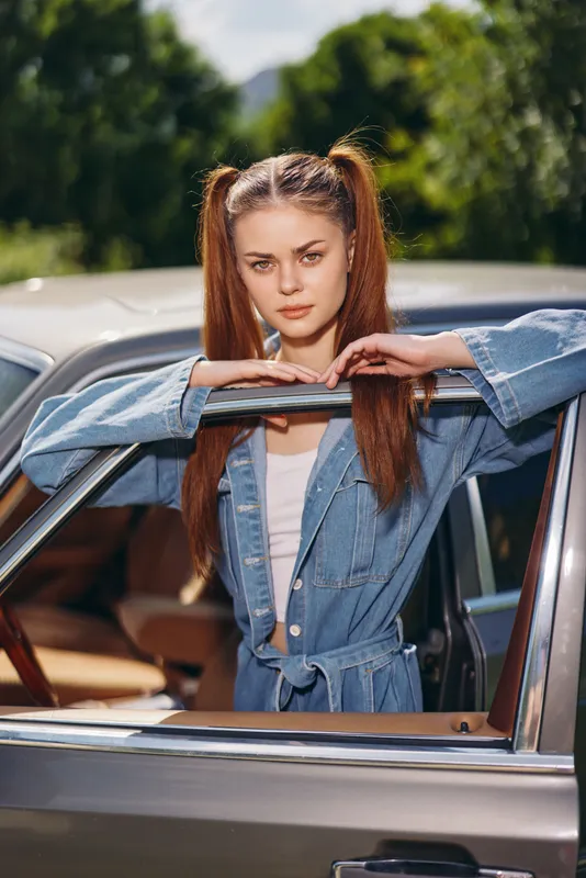 Young woman with long hair and playful pigtails posing in a denim jacket by a classic car in a sunny outdoor setting, capturing a vibrant summer mood.
