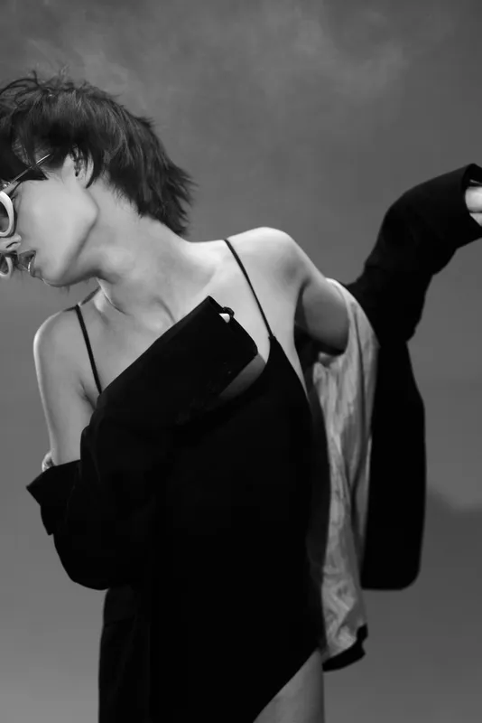 Black and white stylish woman with short hair wearing asymmetric dress posing confidently in studio with minimalistic background.