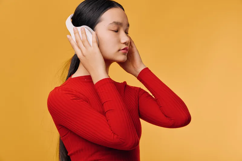 Young woman with long hair listens to music with white headphones wearing red sweater on yellow background. Relaxed female enjoying audio with closed eyes and calm expression.