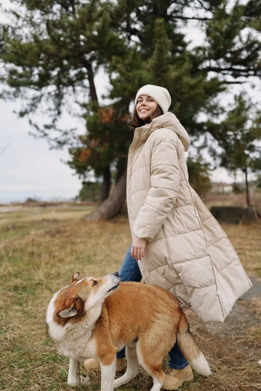Woman in a puffy beige coat and beanie walking with a friendly corgi in a park on a cool day, trees in the background, candid outdoor moment.
