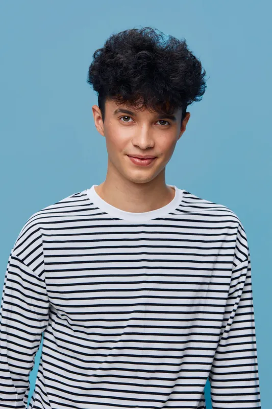 young man with curly hair smiling confidently in casual striped shirt against a blue studio background, stylish and friendly expression.
