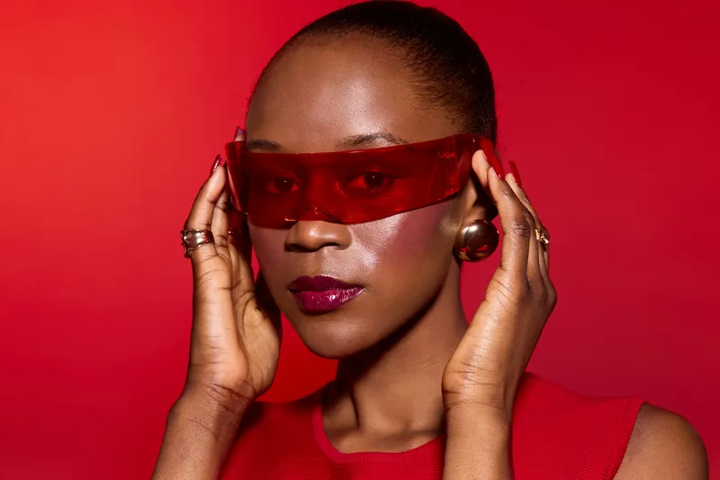 Close-up of a confident black woman with dark skin wearing red transparent sunglasses and a red outfit. Studio portrait on red background with bold fashion and beauty concept.