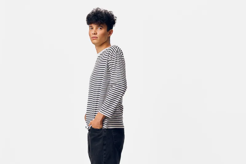 young man wearing striped shirt and black jeans standing sideways with hands in pockets against white background, casual style and thoughtful expression