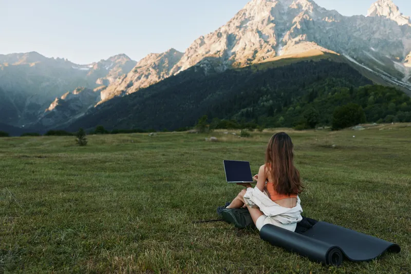 A person sits on a yoga mat in a green meadow with a laptop, surrounded by mountains, enjoying nature and remote work in a peaceful outdoor setting.