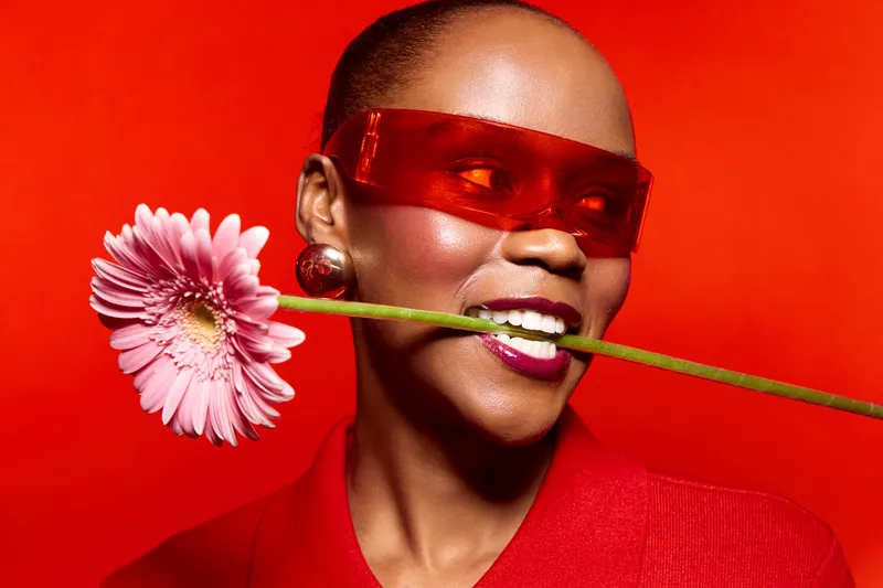 Smiling black woman with dark skin wearing red sunglasses and red clothing holds pink flower stem in mouth against bright red background. Concept of fashion, beauty, and vibrant color contrast.