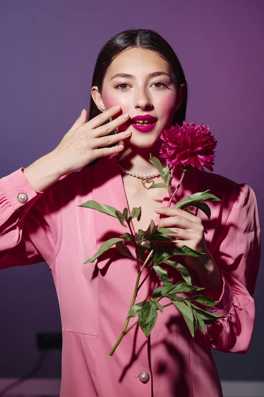 Young woman in fashion pink blazer holding a bright pink flower near her face with vibrant makeup. Creative beauty concept, studio lighting on a purple background.