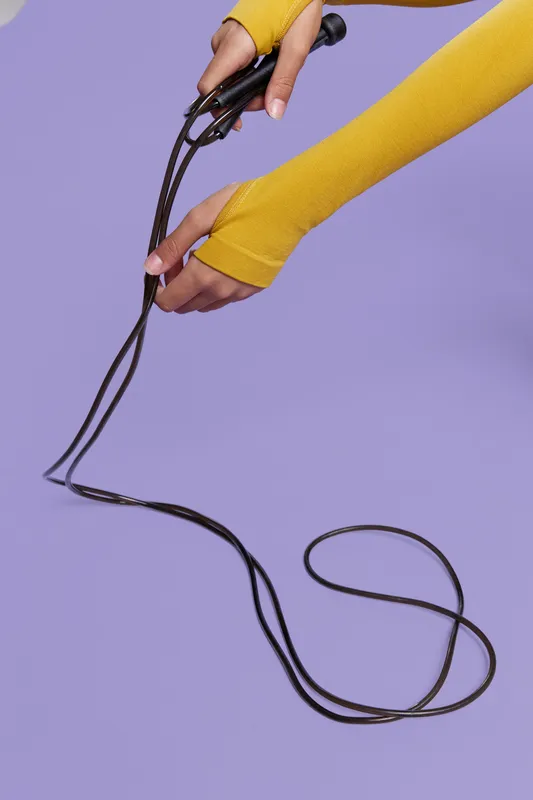 Woman sport fitness hands holding black jump rope, wearing yellow fingerless gloves, against purple background, showcasing active lifestyle equipment and preparation for exercise.