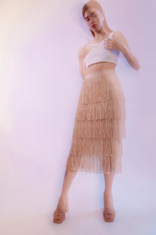 Young woman in a white crop top and beige fringe skirt poses confidently in soft lighting. Concept of woman beauty fashion and modern style with elegant mood.