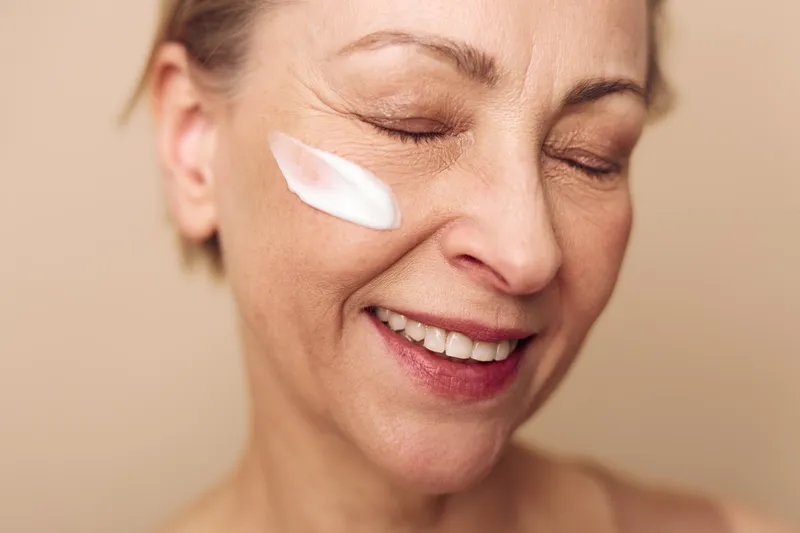 Close-up of mature woman with natural skin smiling with eyes closed while applying moisturizing cream on cheek. Concept of skincare, anti-aging, beauty, and facial care routine.