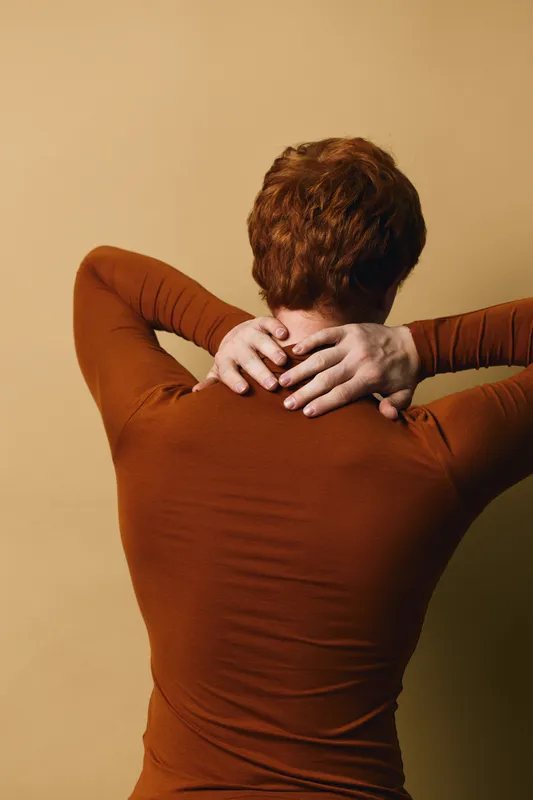 Person with red hair wearing brown sweater standing with back to camera and hands on neck against beige background for wellness and relaxation concept.