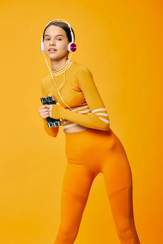 Fit young woman in orange athletic set holds small dumbbells and wears pink headphones against vivid orange background, posing for a dynamic fitness and sport lifestyle portrait.