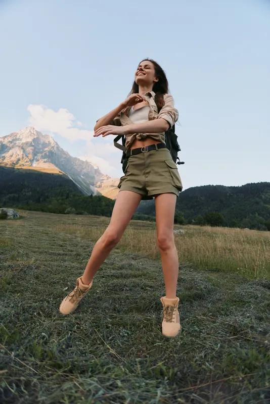 Young woman with wavy hair leaps across a grassy meadow, wearing khaki shorts and a light beige top, exuding freedom and adventure in a bright outdoor scene.