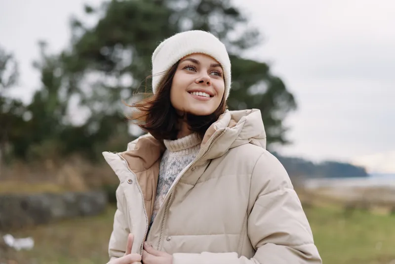 Woman outdoors in a warm beige coat and knit hat, smiling on a chilly day by the coast, with trees and open field in the background, casual seasonal fashion and relaxed mood.