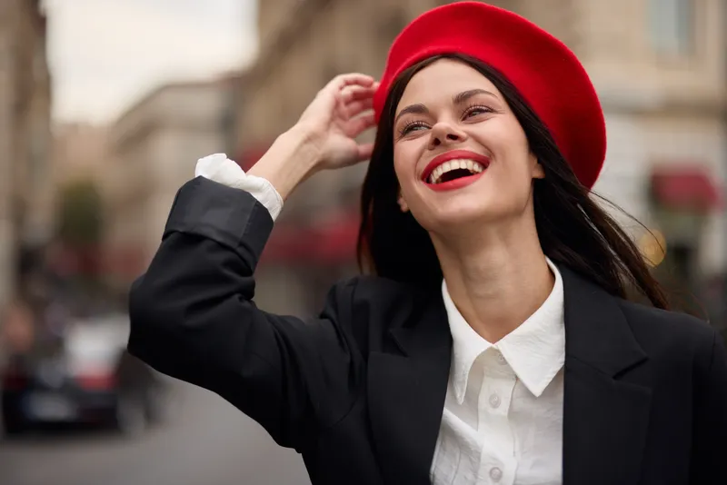 Модный женский портрет: улыбающаяся девушка с зубами стоит на улице на фоне города, одетая в стильную одежду, с красными губами и красным беретом