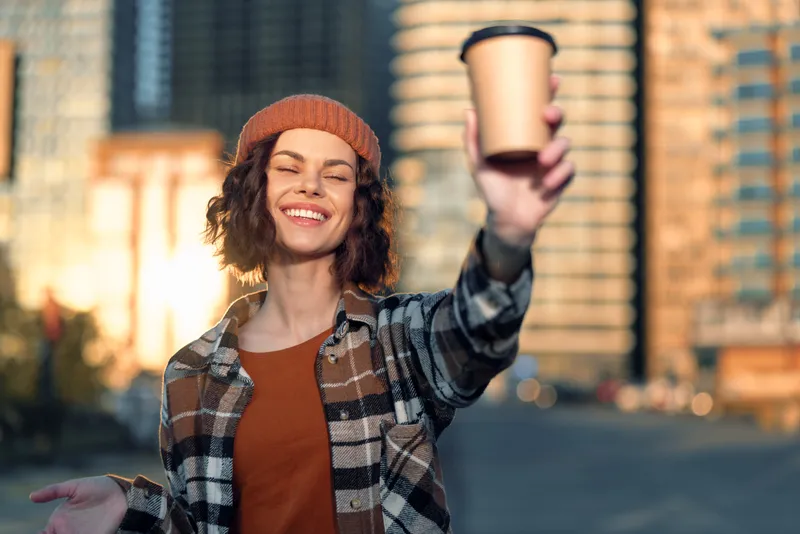 Woman holding coffee cup, smile in city street with beanie and sunlight, a candid lifestyle portrait capturing golden hour glow, authenticity, mindful living and emotional storytelling.