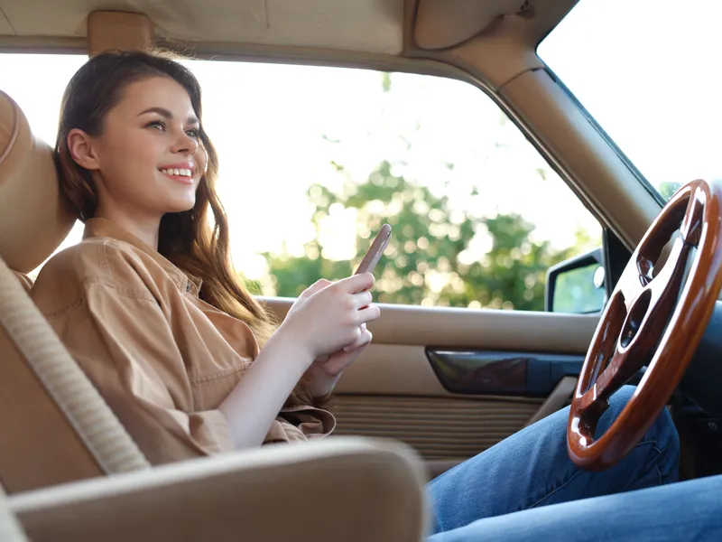 Young woman enjoying a sunny day in a vintage car, smiling and using her smartphone, embodying carefree summer vibes with a warm color palette.