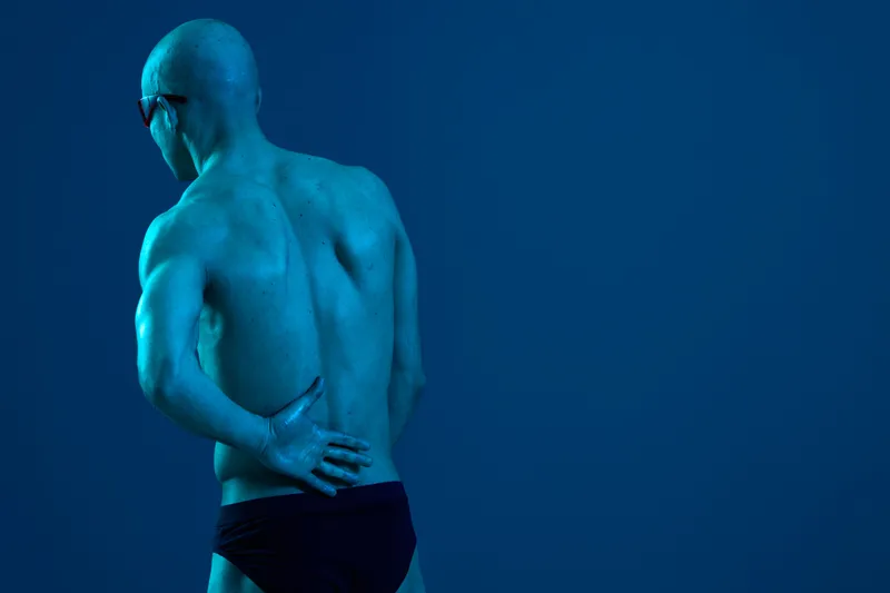 Athletic man experiencing back pain, dressed in swimwear, showcasing a fit physique. The image has a deep blue background, emphasizing the emotion of discomfort.