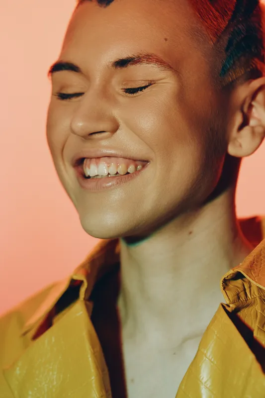 Smiling woman with short hair wearing a yellow jacket, eyes closed and expressing happiness, soft warm lighting on peach background with a joyful mood.