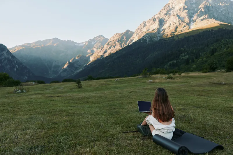 A person sits on a grassy hillside with a laptop, facing a towering mountain range under clear skies, creating a peaceful remote work or nature retreat atmosphere.
