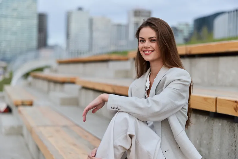 young woman in light suit smiling sitting on urban wooden benches with modern cityscape background business fashion concept