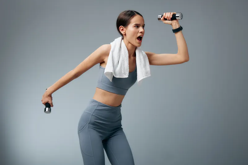 Energetic woman in athletic wear lifts dumbbells with a towel over her shoulder, mid-motion during a powerful workout, showcasing fitness training, strength and concentration in a studio setting.
