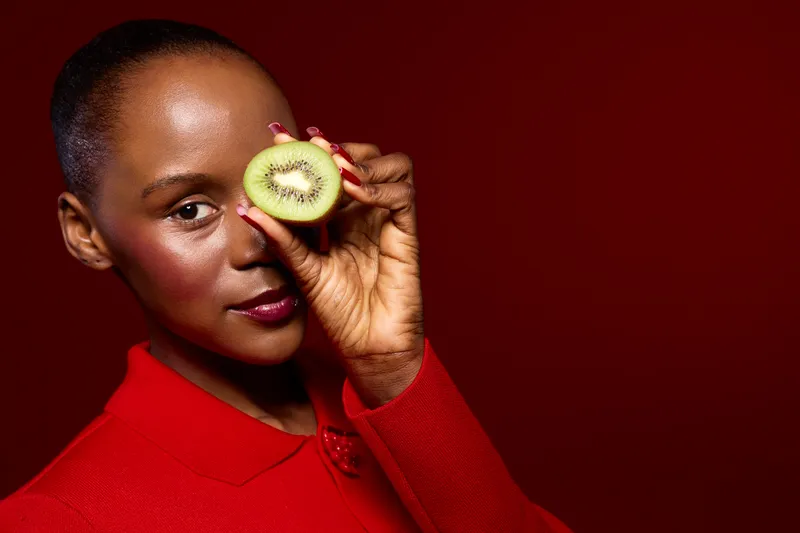 Portrait of dark skinned African woman in red outfit holding half of kiwi fruit near her eye on red background. Concept of healthy lifestyle, natural beauty, and fresh fruit.