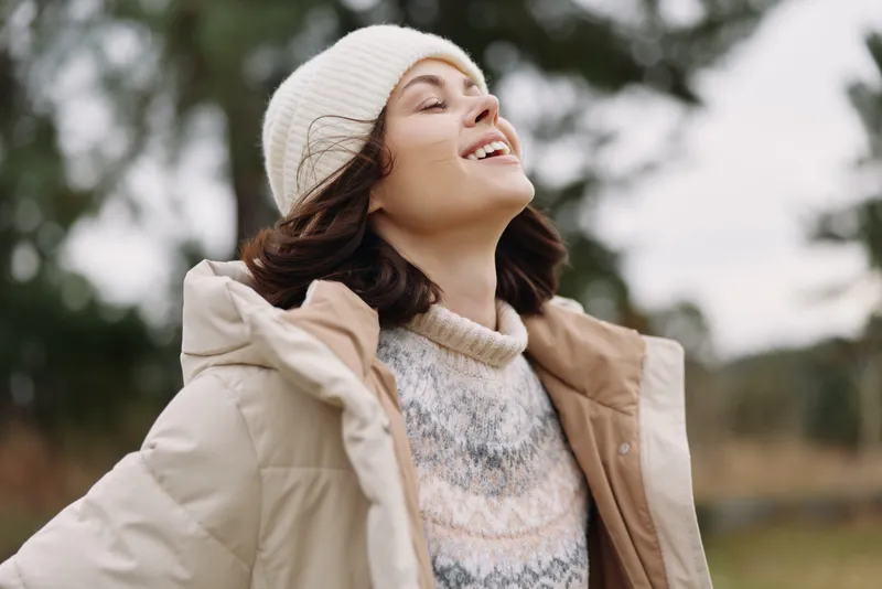 Joyful woman in a beige coat and knit sweater outdoors, enjoying a breezy moment with a beanie, as sunlight and fresh air highlight her relaxed expression in nature.