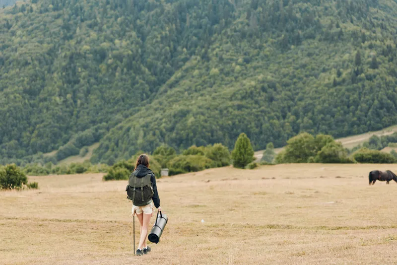 A lone traveler with a backpack walks across a wide meadow, carrying a rolled mat under one arm, with distant hills and trees in a serene outdoor scene.