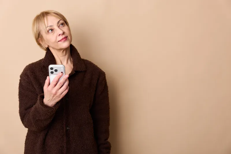 Elderly woman indoors studio portrait, holding smartphone in her hands, smiling softly, looking at camera, beige background, warm light, calm mood today