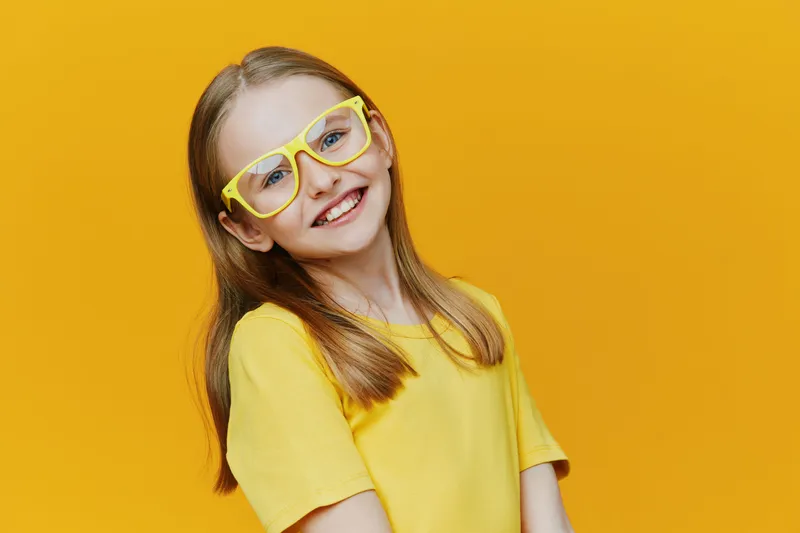 Smiling young girl with glasses on a vibrant yellow background, wearing a bright yellow shirt, showcasing happiness and confidence, perfect for children s fashion or cheerful concepts