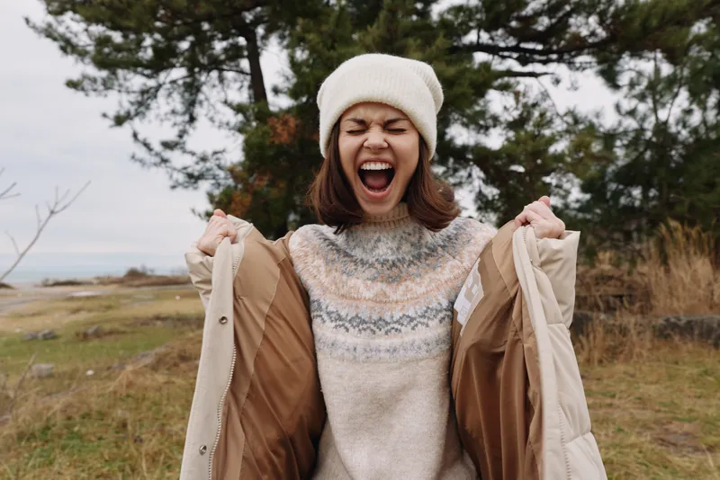 Woman in a cozy sweater and beanie on a windy outdoor day, laughing and pulling open a light coat, framed by an open field and trees in a natural landscape.