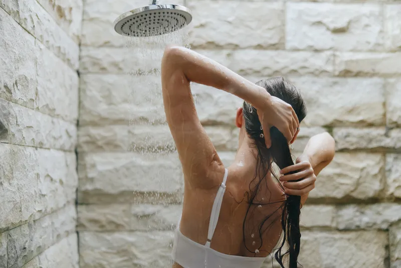 woman washing hair under indoor shower with stone wall background, wet long hair, skin care routine, relaxation moment in bathroom, natural light