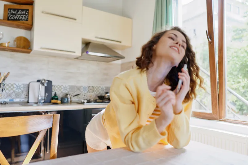 Cheerful woman at home, seated by a sunlit kitchen table, wearing a yellow sweater, talking on a smartphone, relaxed pose, warm tones, candid lifestyle moment indoors. Calm ambient glow.