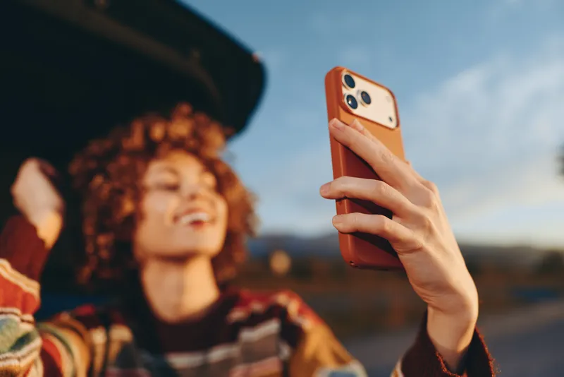 Woman holds smartphone for selfie during outdoor lifestyle moment, hand in focus, curly hair and face softly blurred in background, warm afternoon light, casual street style, urban