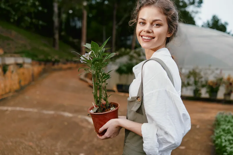 Smiling young woman holding a potted plant in a greenhouse, showcasing gardening enthusiasm and a passion for nature