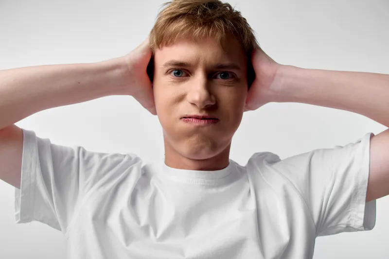 A frustrated man holds his head with both hands, showing discomfort and stress in a studio setting, wearing a white t shirt, conveying tension and mental strain