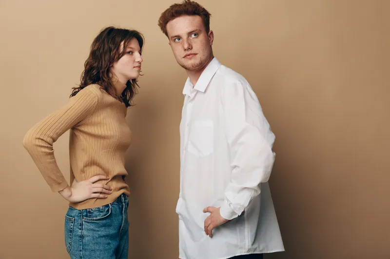 A young woman in a brown sweater confronts a man in a white shirt, both showing serious expressions against a beige background. Concept of conflict and tension.