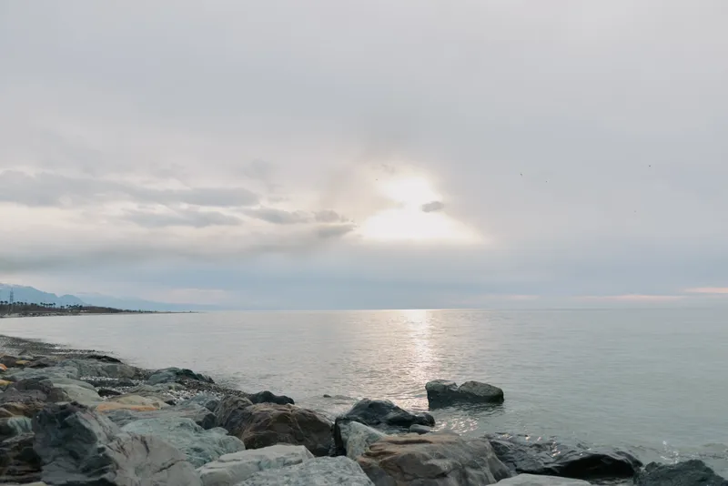 Rocky shore under cloudy sky with calm sea reflecting soft sunlight. Peaceful coastal landscape with gentle waves and distant horizon on a tranquil day by the water.